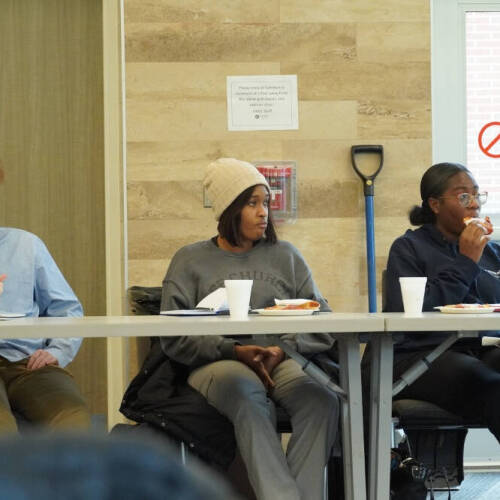 Three people sit at a long table with notebooks, paper cups, and plates of food in front of them. Behind them is a beige wall, a blue-handled shovel, and a door with a "no entry" sign. The setting appears to be a casual meeting or discussion.